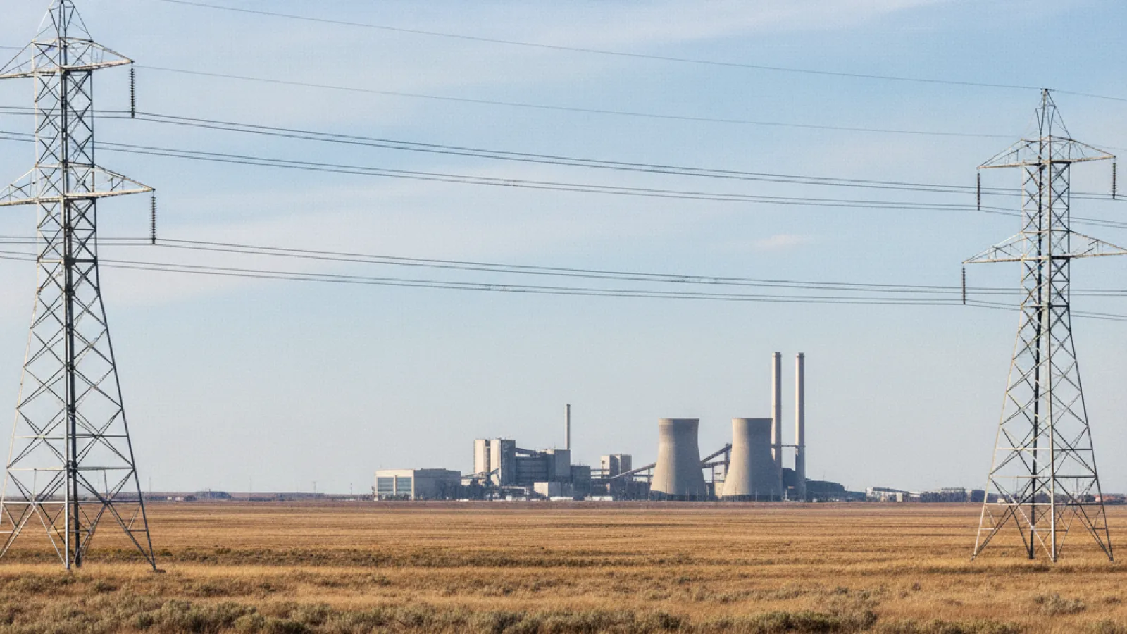 Power transmission towers and lines span a dry, golden field with a power plant, featuring cooling towers and smokestacks, in the distance under a daylight blue sky.